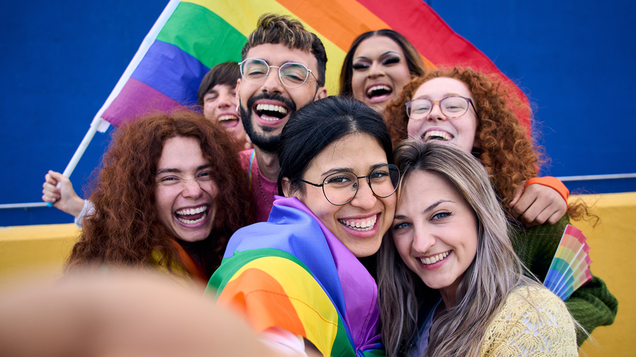 Selfie of a LGBT group of young people celebrating gay pride day holding rainbow flag together. Homosexual community smiling and taking cheerful self portrait. Lesbian couple and friends generation z