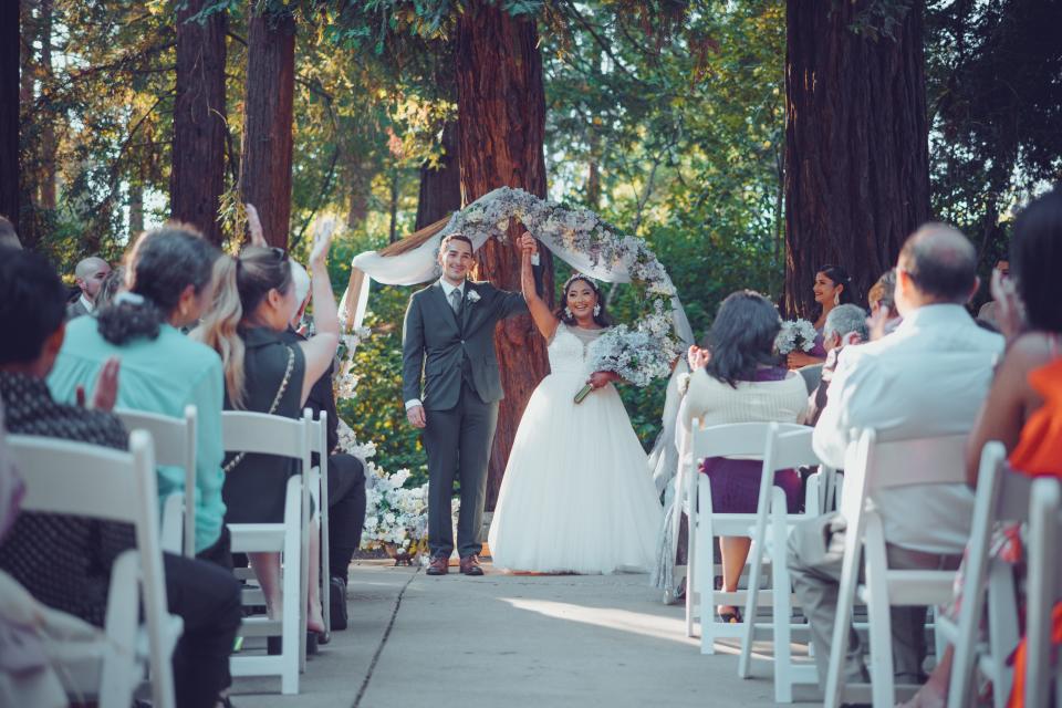 The couple have completed their Christian wedding ceremony and have their hands up as their guests clap. They are outdoors and under a frame of purple flowers. The bride is in a white gown with a purple bouquet.