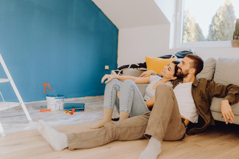 A young couple sits on the floor admiring a freshly painted turquoise wall that they painted together. 