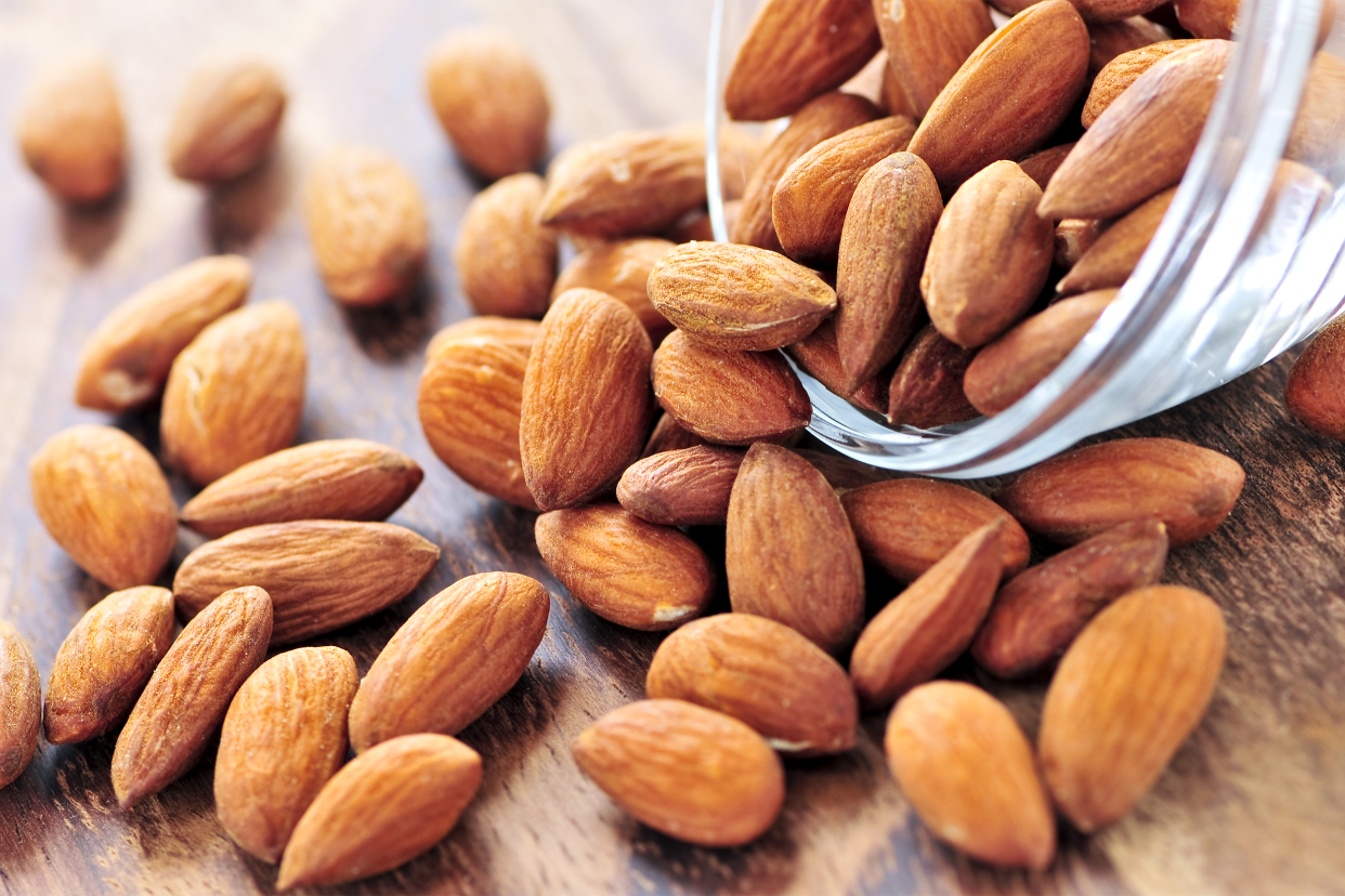 Closeup of almonds being spilled out of a glass bowl on it's side, on a wooden table