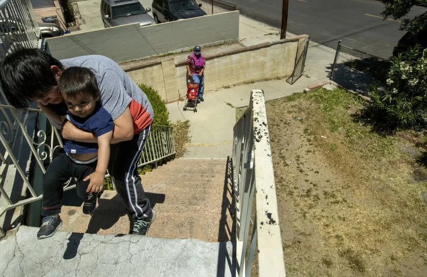 LINCOLN HEIGHTS, CA-MAY 11, 2022: Benita Perez, 55, teaches her grandson Nevaeh Perez, 1 and 1/2, how to walk at their apartment building on Thomas St. in Lincoln Heights. Area at right, below used to be all grass until a year ago, due to the drought. It's going to be a summer of brown grass and hard choices for Southern California lawn owners facing the Metropolitan Water District's one day a week watering restrictions starting June. 1. (Mel Melcon / Los Angeles Times)
