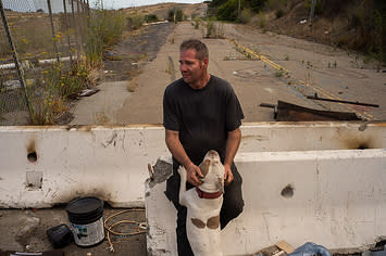 A man sits on a concrete barrier surrounded by debris, while his dog jumps up on him