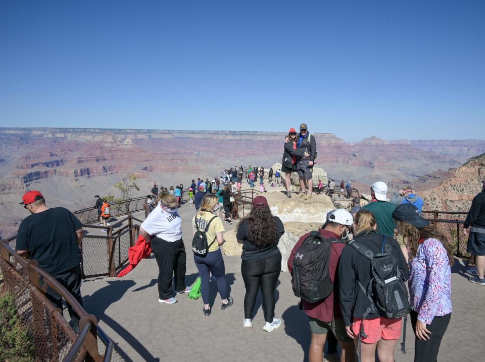 Several groups of people stand in front of the view of Mather Point at the Grand Canyon on a clear day with blue skies.