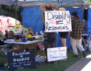 A photo showing several signs referring to disability justice and Palestine's connection to disability justice. One sign says the tent has disability resources, including masks, earplugs and other supplies to help make protesting more accessible for disabled students. 