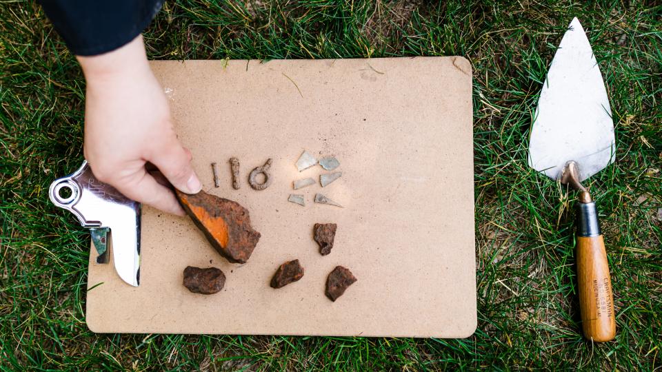 Three iron screws, six flat glass shards and five red brick fragments on brown clipboard. A hand is holding the largest brick fragment. The clipboard lays on grass and a trowel is laying on the on the right side of the clipboard.