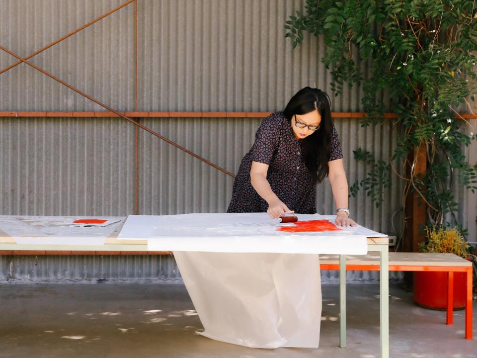 A woman leans over a table painting red paint on paper.