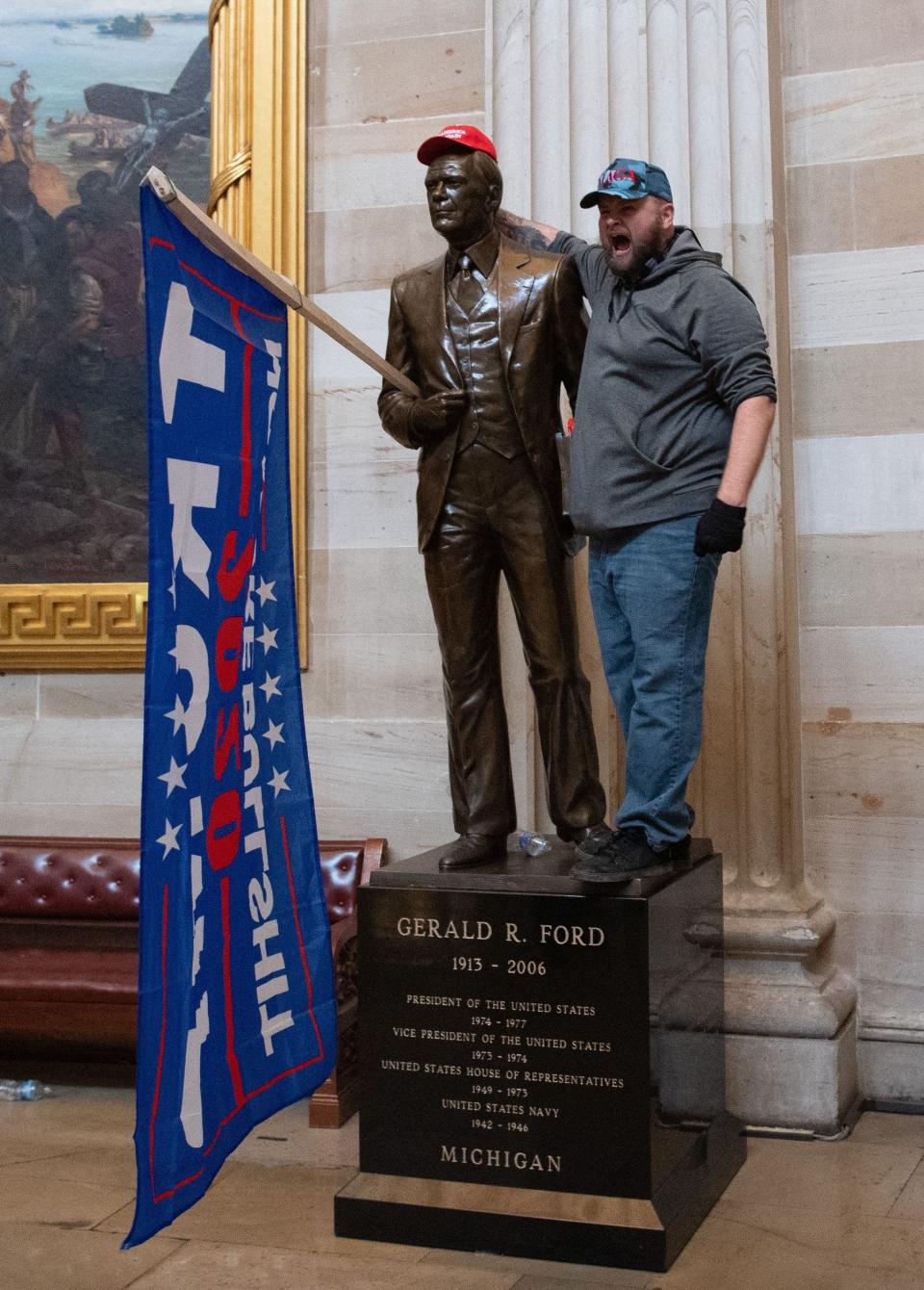 A pro-Trump supporter puts a MAGA hat and a Trump flag in the arm of a statue of Gerald Ford