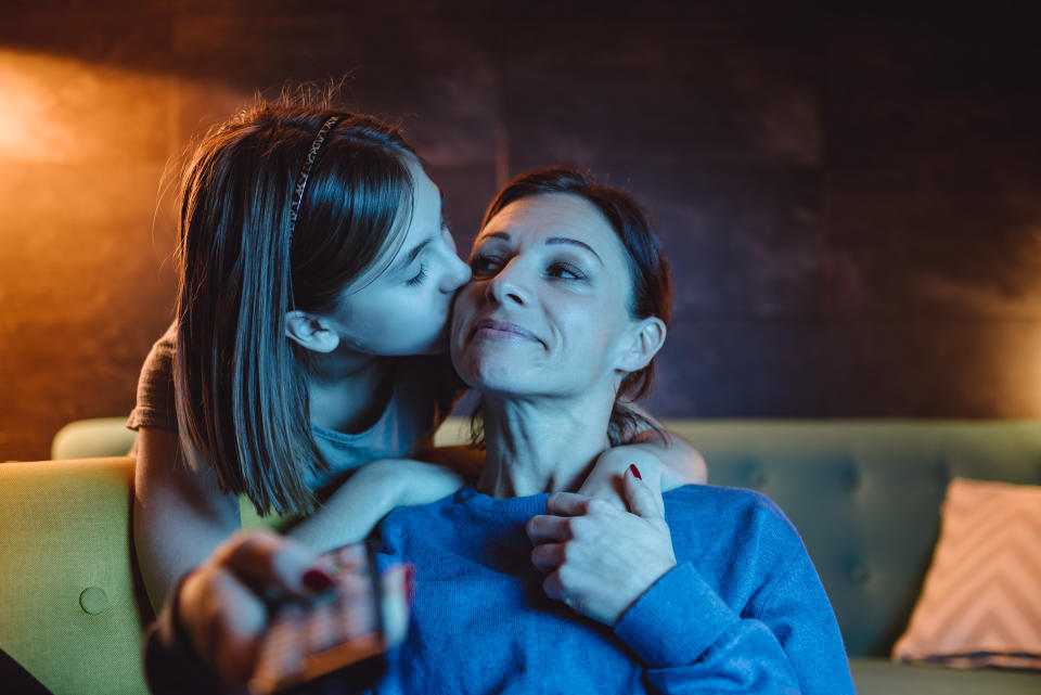 Mother sitting on sofa and watching TV late at night while daughter giving her goodnight kiss
