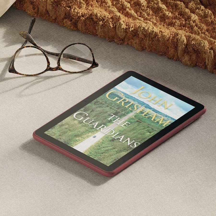 An Amazon tablet with a John Grisham book cover, next to a pair of glasses and a blanket. 