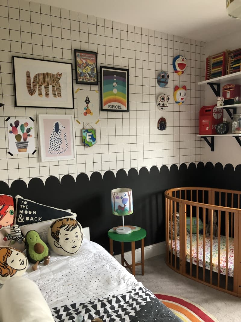 graphic grid on upper half of wall, scallop wall divide, black bottom half of wall, oval baby crib with wooden slates, kids bed, avocado throw pillow, face throw pillows, gallery wall, floating shelves, small green side table, illustrated lamp shade, black and white comforter