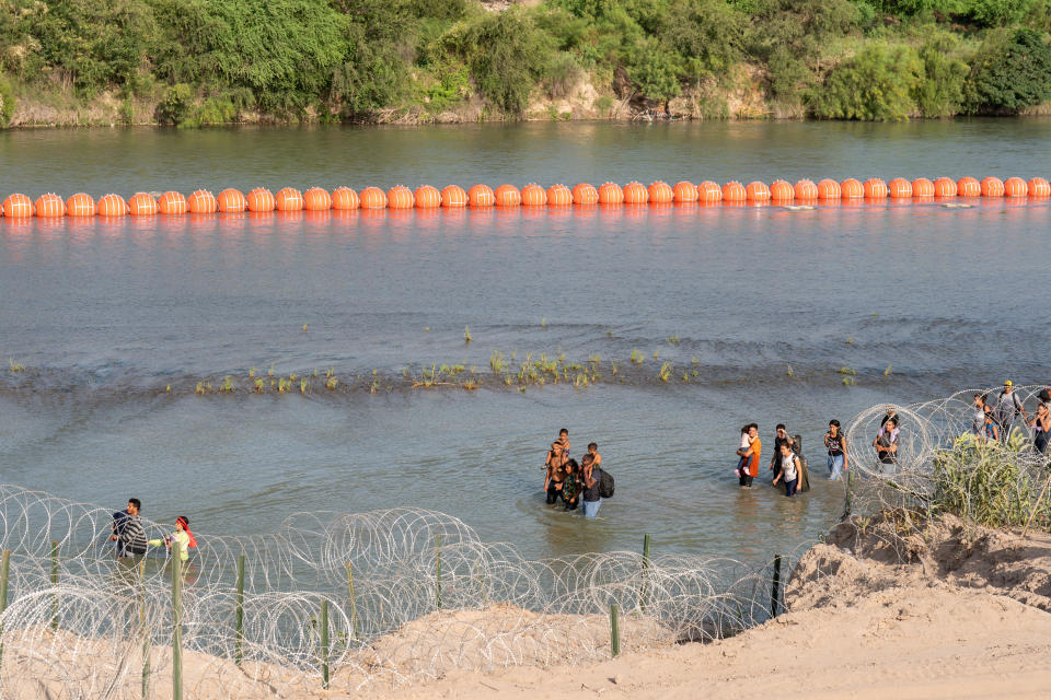 Migrants walk by a string of buoys placed on the water along the Rio Grande border with Mexico.