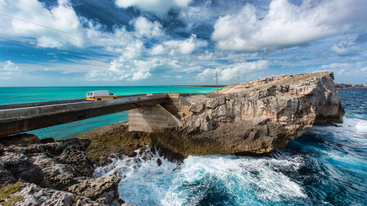  A van crosses a bridge, with turquoise water one one side and waves breaking on rocks on the other, at the Glass Window bridge, Eleuthera island, Bahamas. 