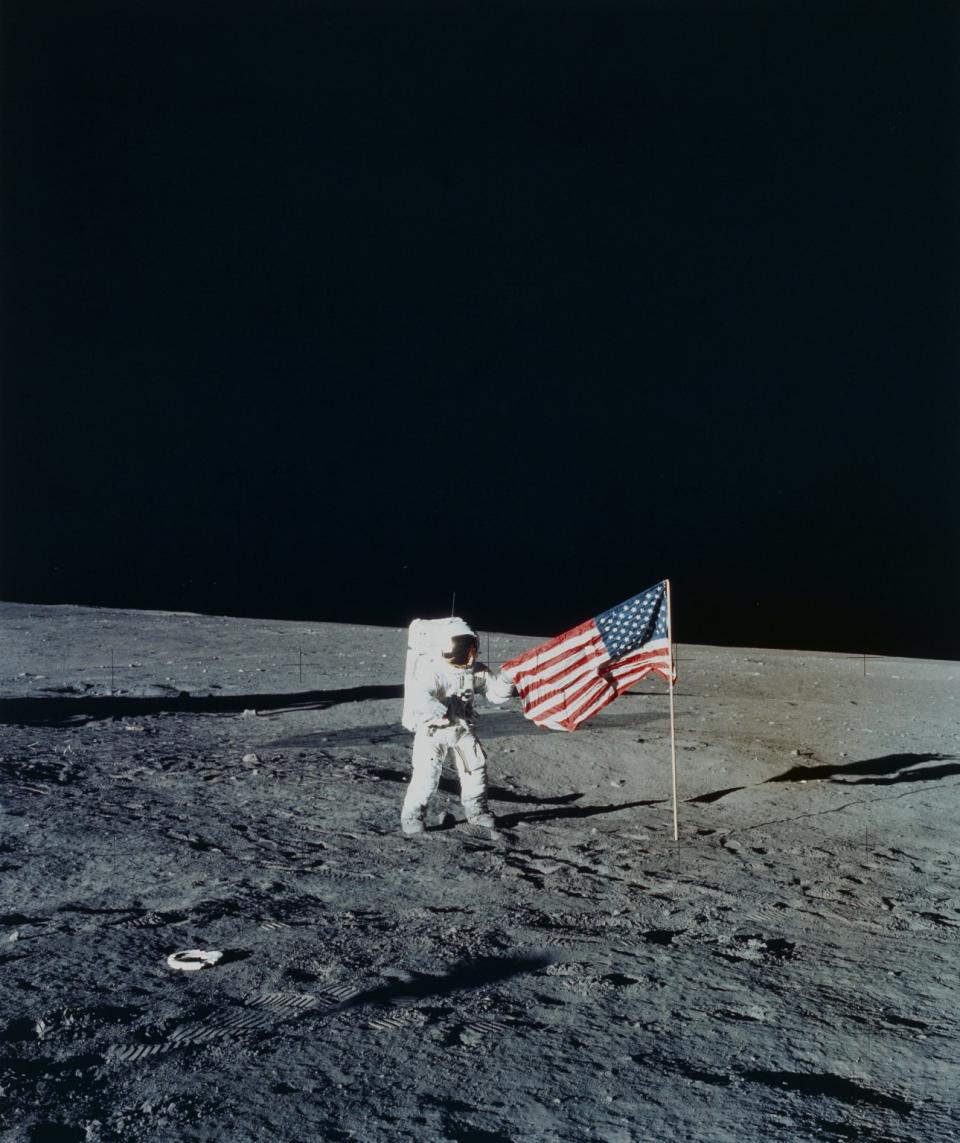 An astronaut in a bulky white spacesuit plants an American flag on the dusty gray surface of the moon