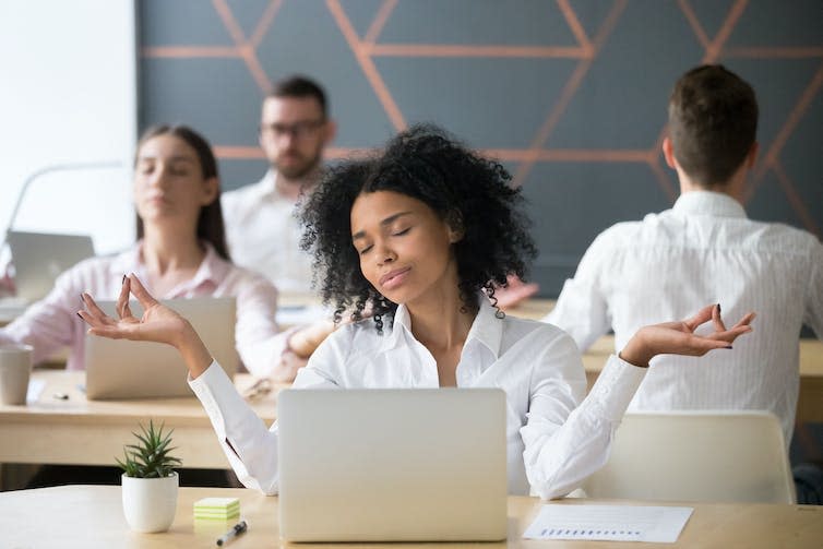 Woman meditating at desk, colleagues in the background, calm, yoga exercises, feeling zen, stress-free, corporate meditation, employee wellbeing.