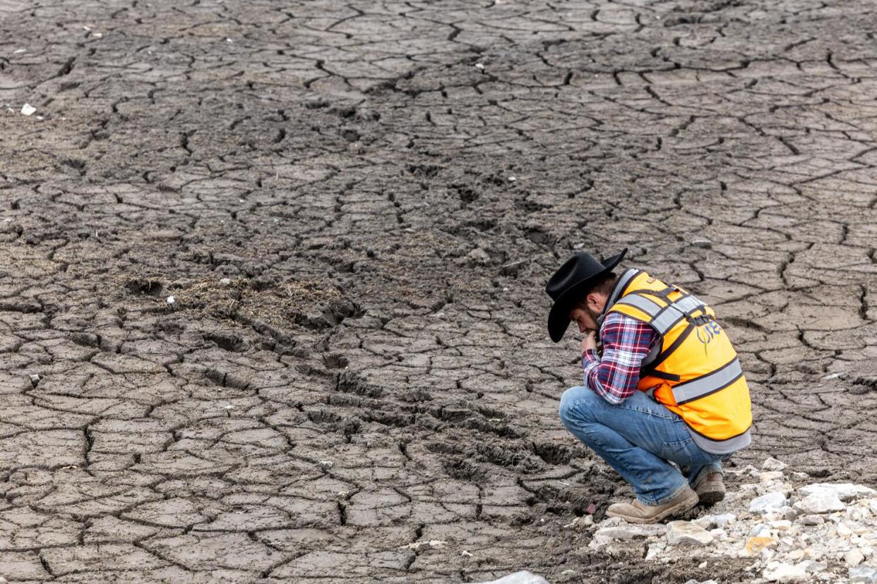 A man in a yellow safety vest squats on a dry spot next to footprints in an expanse of cracked mud, looking down