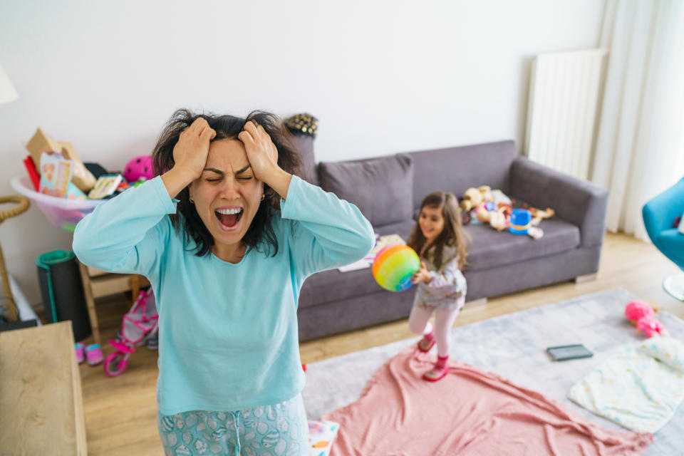 A tired mother in pajamas screams in frustration while holding her head in a messy living room, with a young child playing nearby