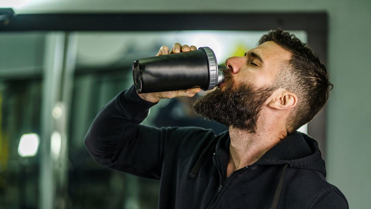 caesin protein powder front view portrait of young caucasian man athlete in black hoodie male standing in the gym holding protein supplement shaker supplementation in training waist up black hair and beard copy space drink