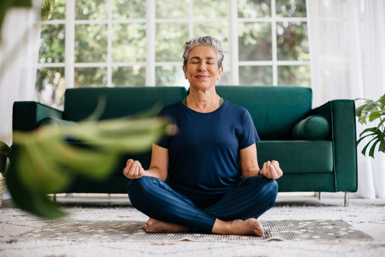 Senior woman meditating in lotus position at home, sitting on the floor in fitness clothing. Mature woman doing a breathing workout to achieve relaxation, peace and mindfulness.