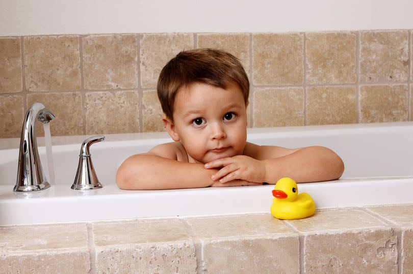 a beautiful toddler boy stares out of the bathtub with large brown eyes, chubby arms folded and rubber ducky by his side