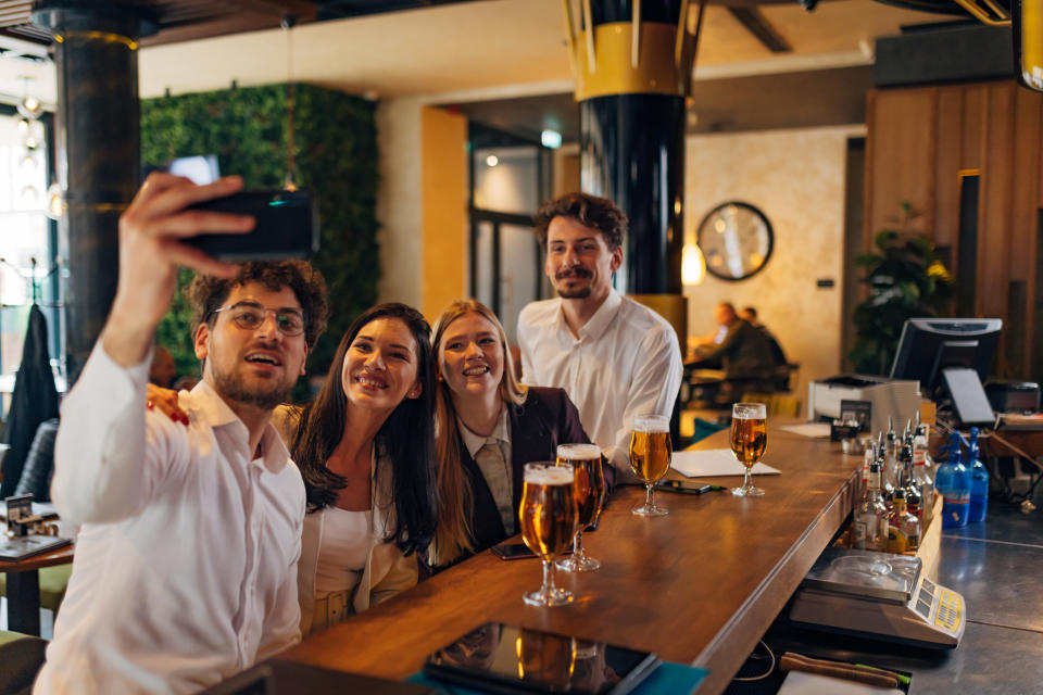 Four colleagues take a selfie at a bar, expressing camaraderie with drinks on the table