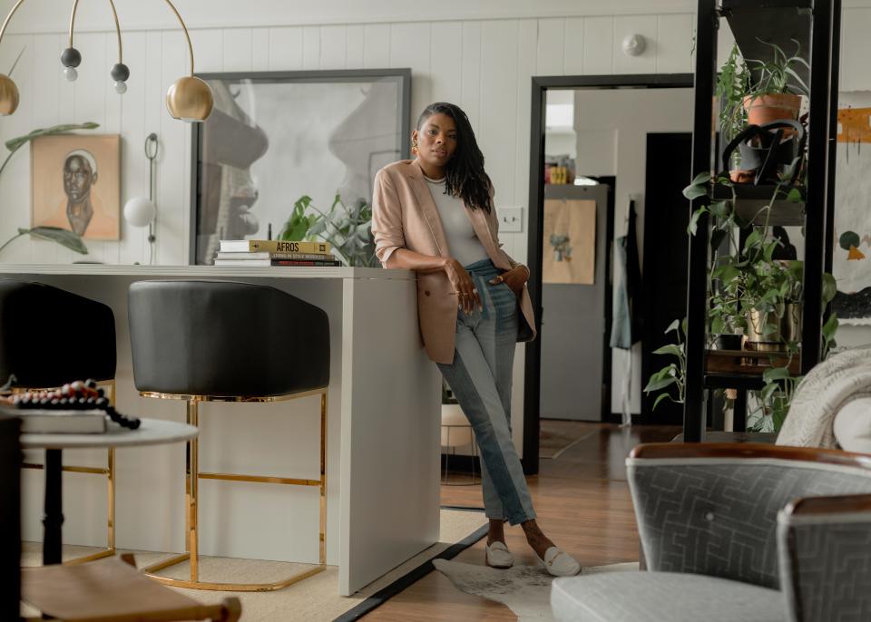 Interior designer Carmeon Hamilton leans on a counter in her home. She's wearing a blush pink blazer over a white shirt with blue jeans and white loafers. Her home is beautifully decorated with plants, paintings, and geometric furniture and fixtures.