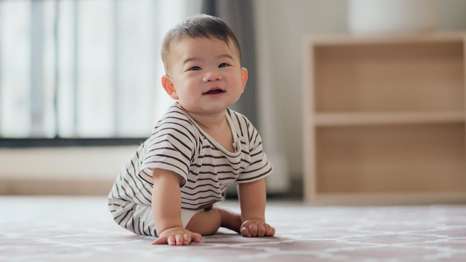 A smiling baby, dressed in a striped onesie, is sitting on a patterned rug inside a home