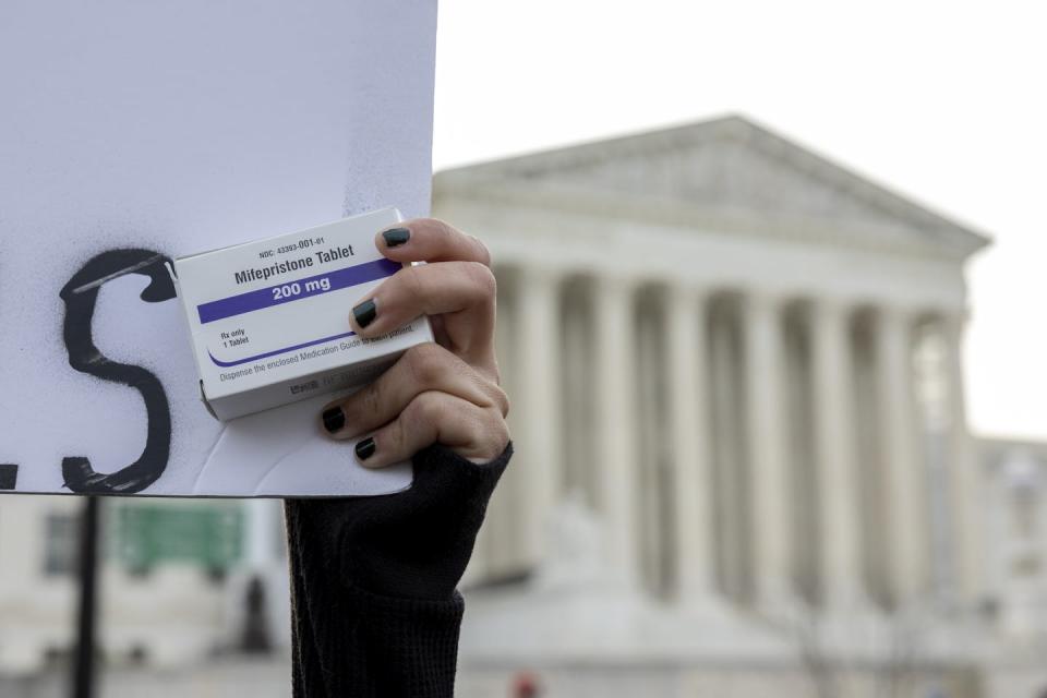 A hand with painted nails clutches a small package of medicine in front of a marble building with tall pillars.