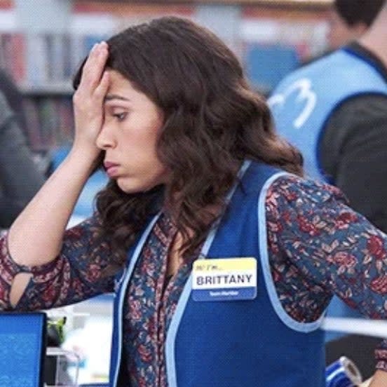 Brittany in a patterned shirt and blue vest with a name tag looks stressed, resting her head in her hand while working at a store counter