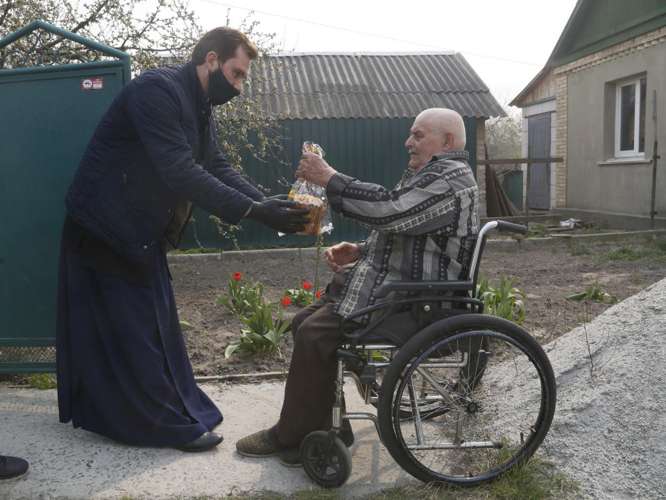 Priest of Ukrainian Orthodox Church Nazariy, wearing a face mask to protect against coronavirus, presents an elderly disabled man with an Easter cake near his house in the village of Nove close to capital Kyiv, Ukraine, Saturday, April, 18, 2020. All the Ukrainian churches have been closed for people because of COVID-19 outbreak, and believers wait for the priest right near their houses. For Orthodox Christians, this is normally a time of reflection, communal mourning and joyful release, of centuries-old ceremonies steeped in symbolism and tradition. But this year, Easter - by far the most significant religious holiday for the world's roughly 300 million Orthodox - has essentially been cancelled. (AP Photo/Efrem Lukatsky)