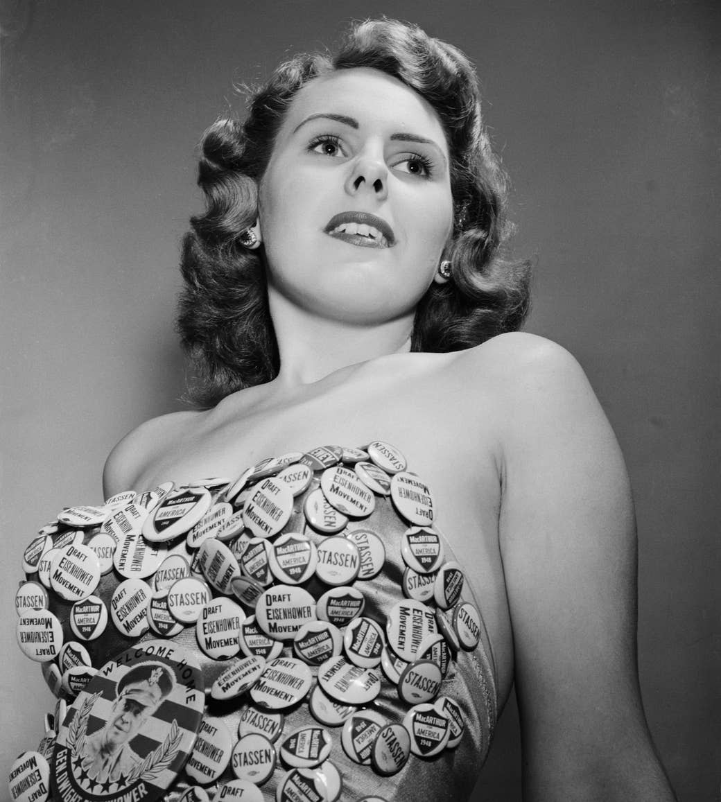 A woman looking into the distance over the camera wears a multitude of campaign pins fixed to her bathing suit that read "draft Eisenhower movement" and the name "Stassen"