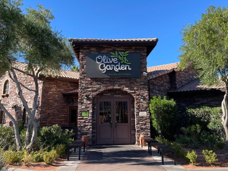 Exterior of Olive Garden. The brick building with an "Olive Garden sign" is surrounded by trees and a bright blue sky