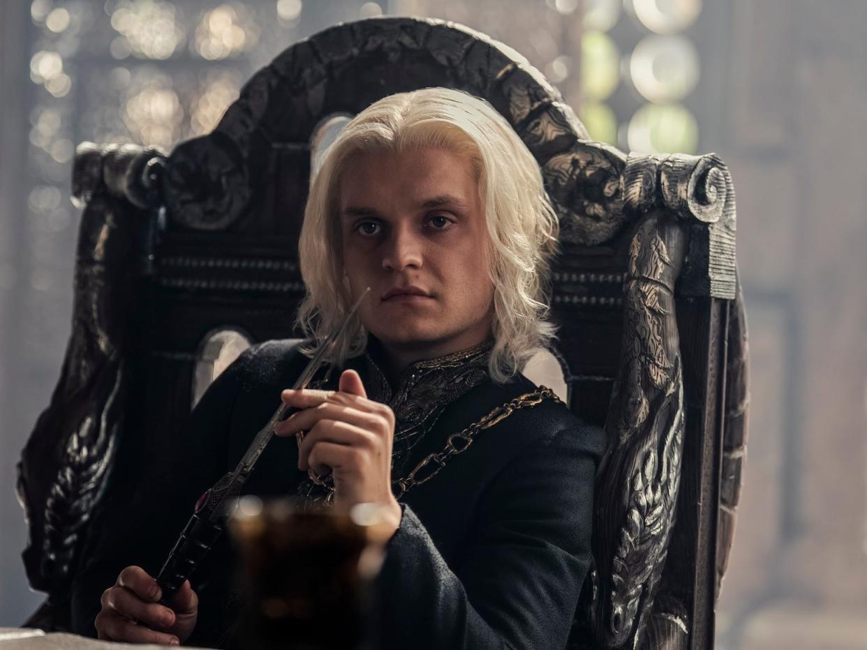 tom glynn-carney as aegon targaryen in house of the dragon. he's sitting in a high backed chair, playing with a steel dagger, and looking across a table