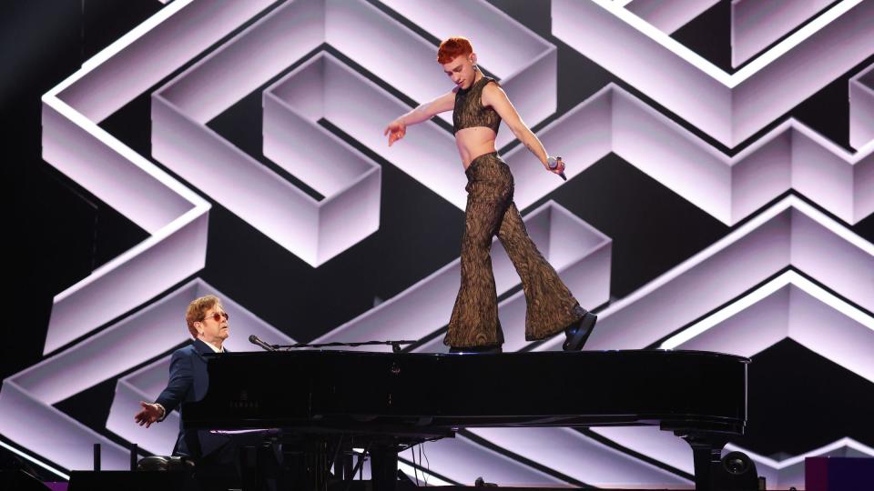 Olly Alexander standing on top of a piano holding a microphone whilst Elton John plays the piano