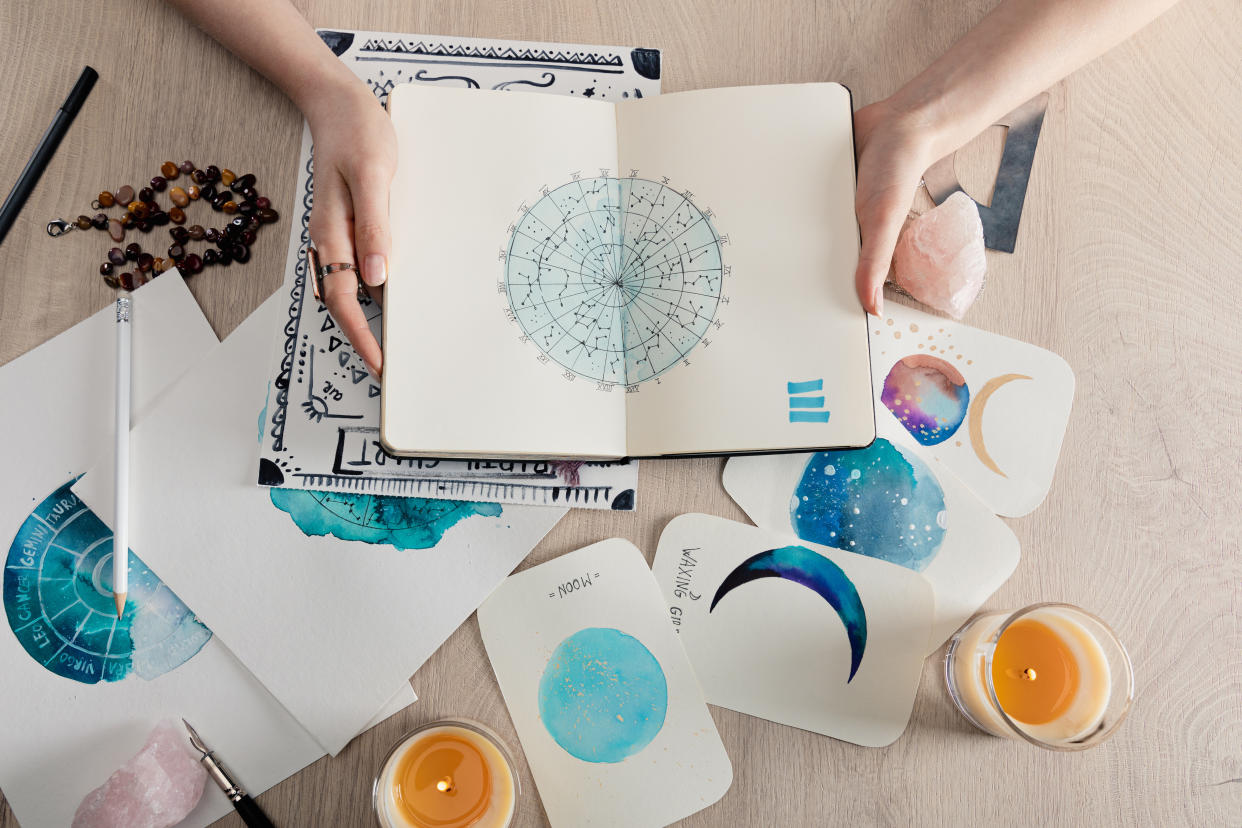 Top view of astrologer holding notebook with watercolor drawings and star signs on cards on table