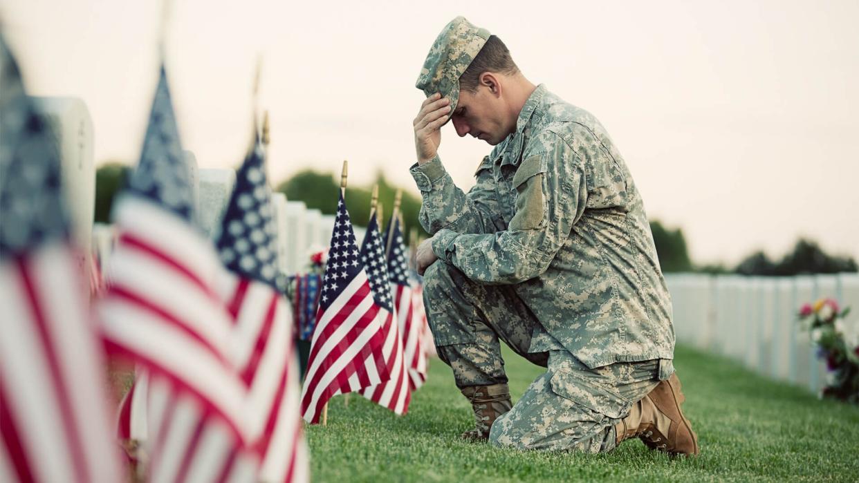 A soldier in ACU fatigues kneels in front of a grave with several American flags in front of it.