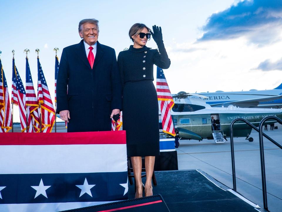 Donald and Melania in a black skirt suit and sunglasses standing next to American flags with an airplane in the background.