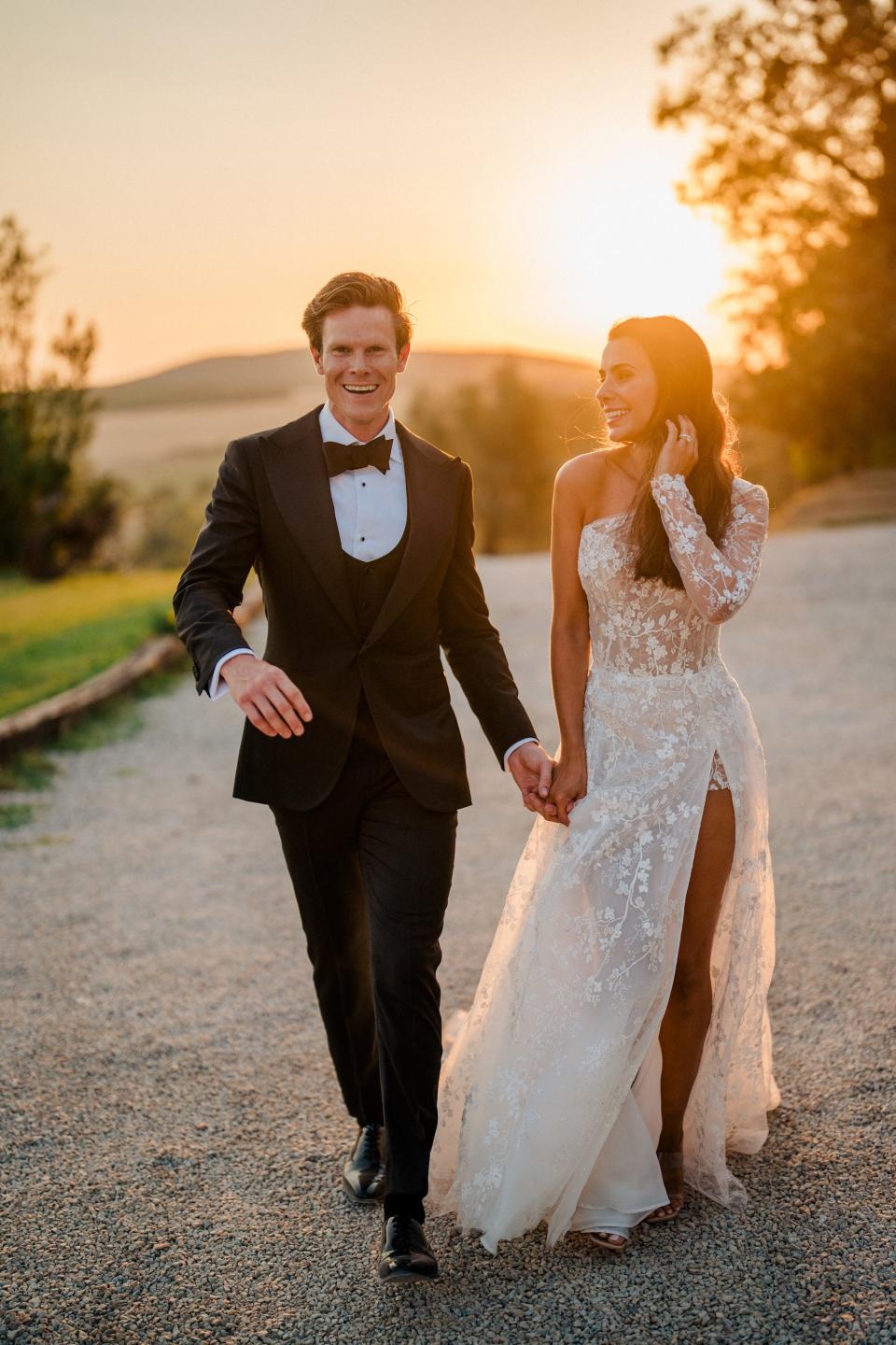 A couple walks down a road in their wedding attire at sunset.