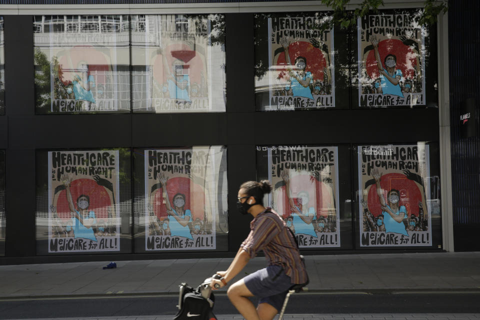 A man wearing a face mask cycles past a coronavirus related artwork displayed on screens in the window of the Flannels clothing store on Oxford Street, in central London, Thursday, May 21, 2020. (AP Photo/Matt Dunham)