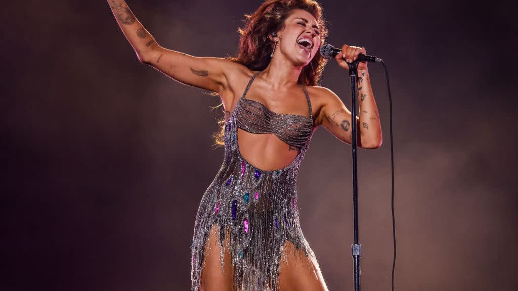 miley cyrus sings into a microphone that she holds on a stand with one hand, she reaches her other arm outward, cyrus wears a beaded strapless body suit with a fringe style skirt