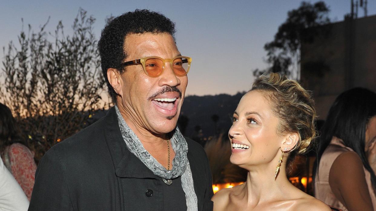 lionel richie and nicole richie stand together at an event, lionel is smiling as he speaks and has one arm around nicole who is looking at lionel and smiling