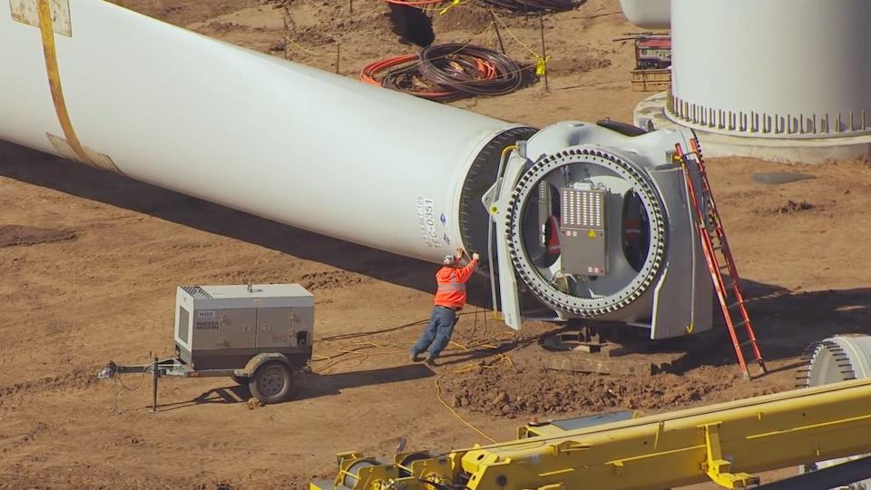 A construction worker looks tiny relative to a wind turbine, which is laid flat on the ground receiving maintenance.