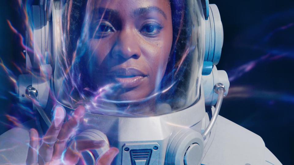 Still from The Marvels (2023) movie. Monica Rambeau gets in trouble. Close up of a woman's face whilst she is wearing a white astronaut spacesuit. She is holding up her right hand and is touching some kind of blue/purple electrical field/film.