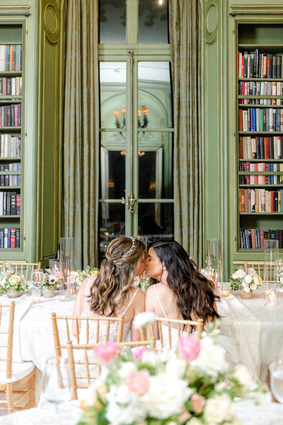 Two brides kiss in front of a green backdrop.