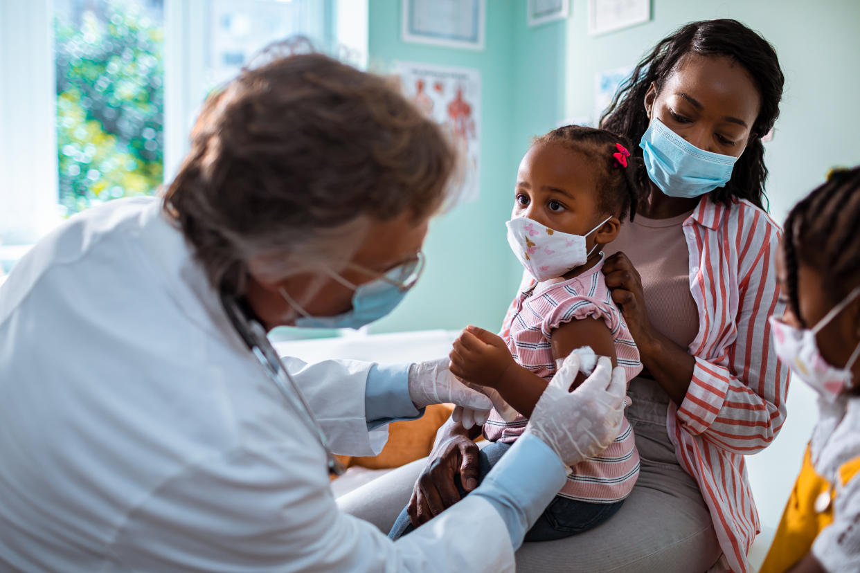 A woman holds her daughter in her lap as a health care worker swabs her upper arm while another girl looks on, all wearing face masks