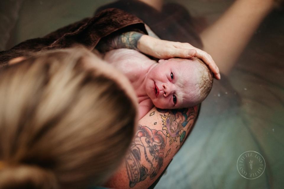 Newborn held by a tattooed parent post water birth, another person's head in the foreground