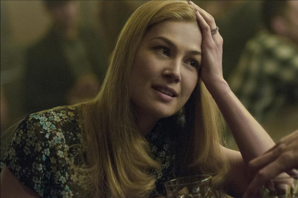 Rosamund Pike rests her head on her hand while sitting at a table, wearing a floral-patterned blouse