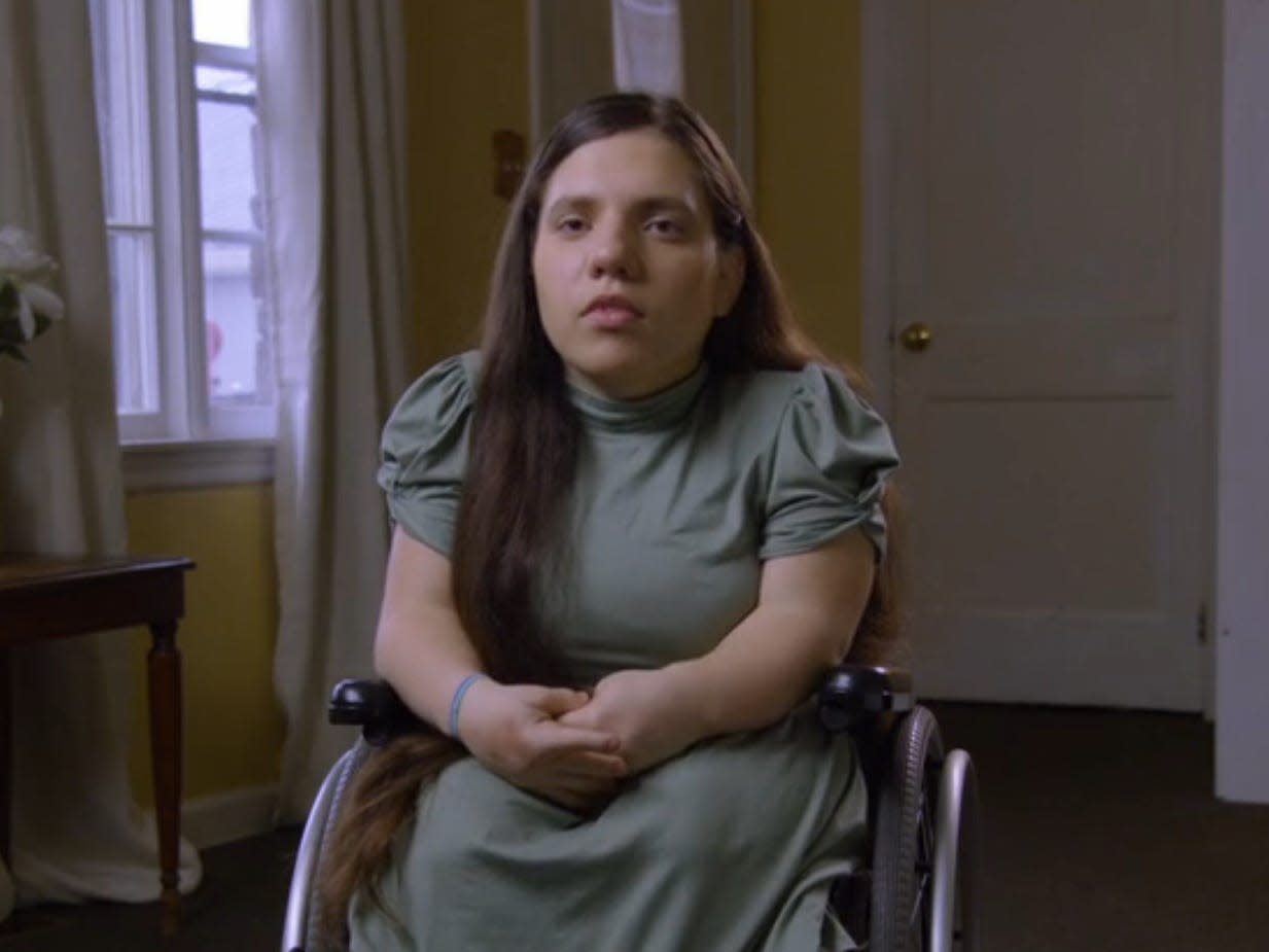 natalia grace barnett, wearing a green dress, her hair long and brown, and sitting in a purple wheelchair in a yellow painted room