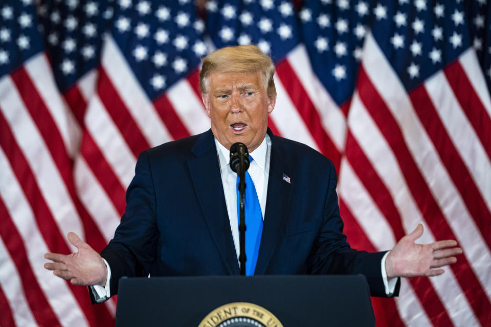 Donald Trump speaks into a microphone at a podium in front of a wall of American flags.