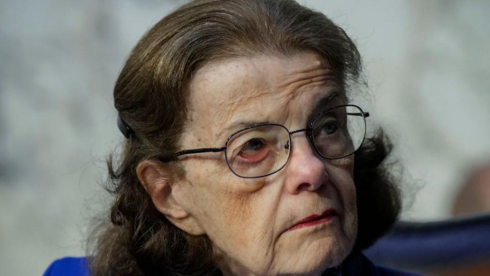 dianne feinstein, wearing a blue outfit and glasses, looks off camera and listens to someone