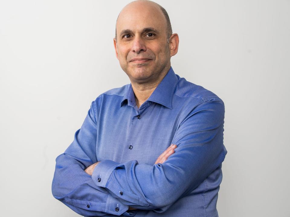 Ron Benegbi, founder and CEO of Uplinq, poses in a blue shirt in front of a grey backdrop with his arms crossed over his chest.